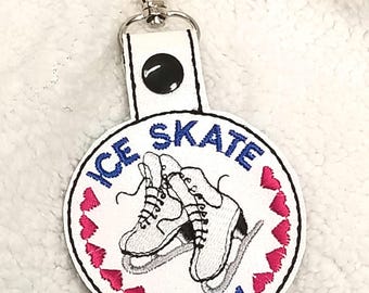 Könnte beinhalten: Weißer runder Schlüsselanhänger mit einem blauen und weißen gestickten Design von Schlittschuhen und dem Text "ICE SKATE MOM" umgeben von rosa Herzen.