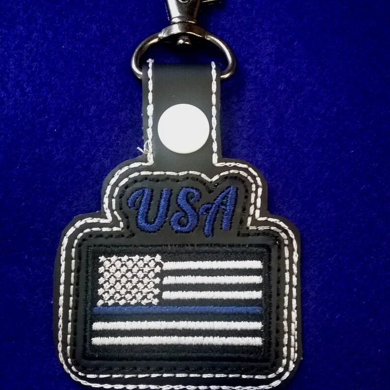 Puede incluir: Llavero en blanco y negro con un dise&ntilde;o de bandera estadounidense cosido. La bandera presenta una franja azul y la palabra "USA" en escritura azul. El llavero tiene un cierre plateado y un c&iacute;rculo blanco.