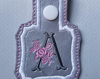 May include: A silver gray key chain with a white button and a pink and black embroidered letter A with floral accents.