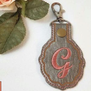 May include: A metallic silver keychain featuring a scalloped edge and a central embroidered pink letter G. The keychain is attached to a silver-toned metal lobster clasp and includes a small circular brass-colored rivet at the top of the tag.