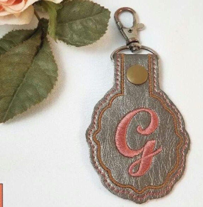 May include: A metallic silver keychain featuring a scalloped edge and a central embroidered pink letter G. The keychain is attached to a silver-toned metal lobster clasp and includes a small circular brass-colored rivet at the top of the tag.