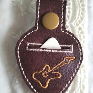 May include: A brown leather guitar pick holder with a white pick inside and a gold button. The holder has a guitar design embroidered on the front.