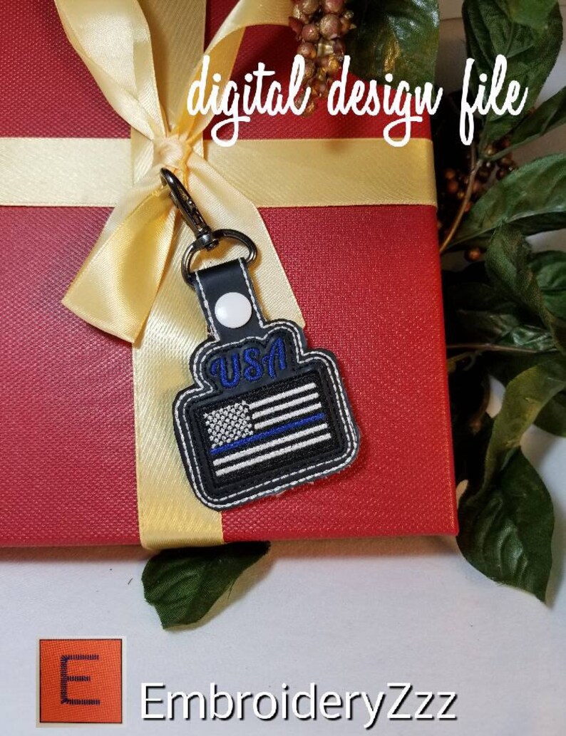 Puede incluir: Un llavero en blanco y negro con un dise&ntilde;o de bandera estadounidense de l&iacute;nea azul, sujeto a una caja de regalo roja con una cinta dorada. El llavero tiene la palabra "USA" en azul. El texto "digital design file" est&aacute; escrito en cursiva blanca.