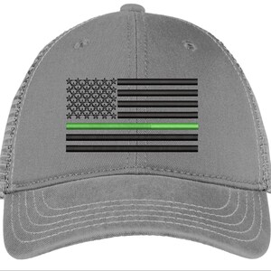 Op de afbeelding: Een grijze truckercap met een Amerikaans vlagontwerp in zwart en groen. De vlag heeft een dunne groene streep in het midden, die de dunne groene lijn voorstelt.