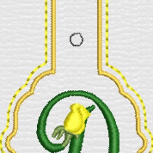 May include: A white tag with a scalloped bottom edge and a rectangular top extension featuring two circular holes. The tag is outlined in yellow stitching and displays a large, green, cursive letter D accented with a small yellow rosebud.