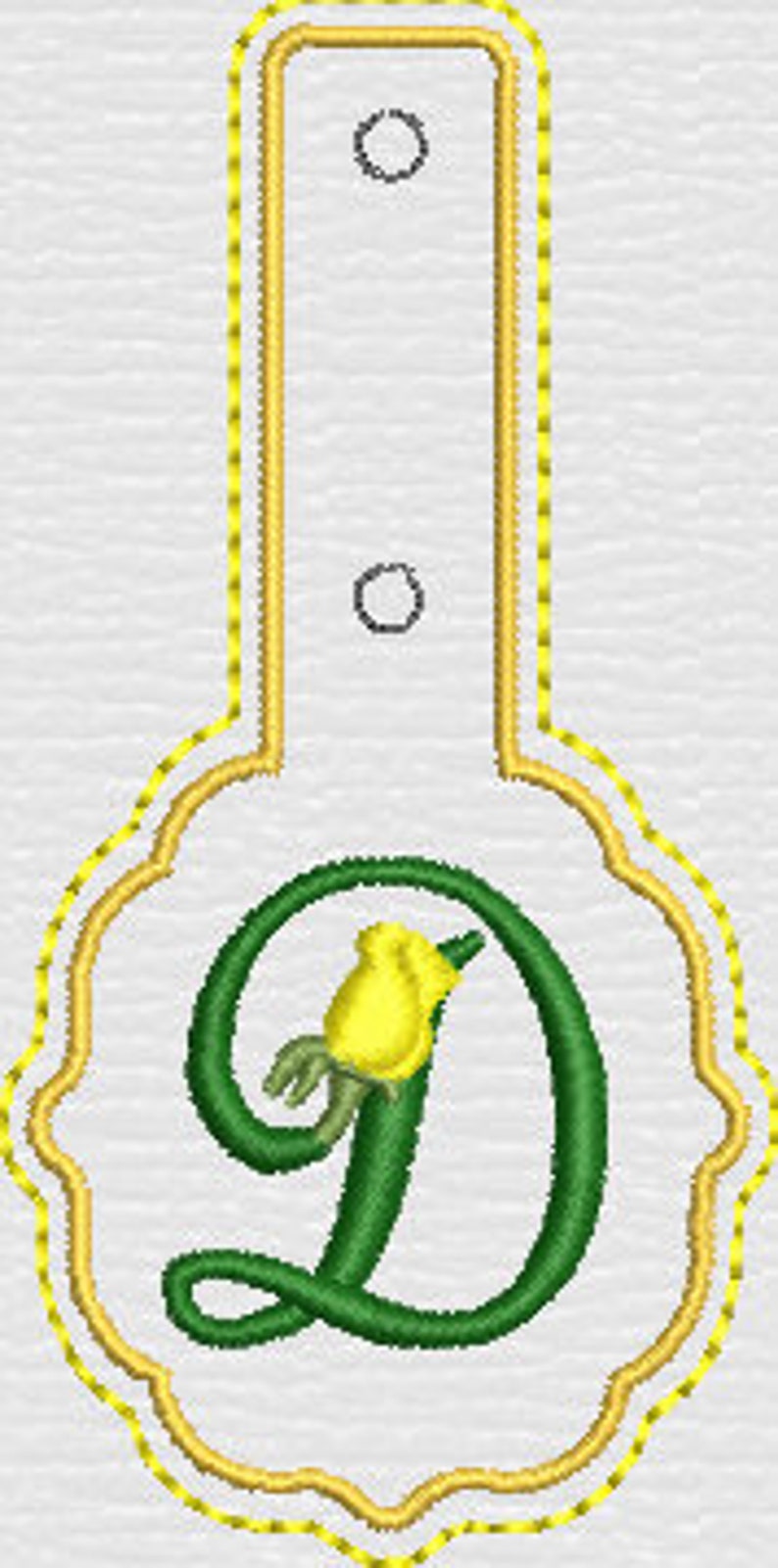 May include: A white tag with a scalloped bottom edge and a rectangular top extension featuring two circular holes. The tag is outlined in yellow stitching and displays a large, green, cursive letter D accented with a small yellow rosebud.