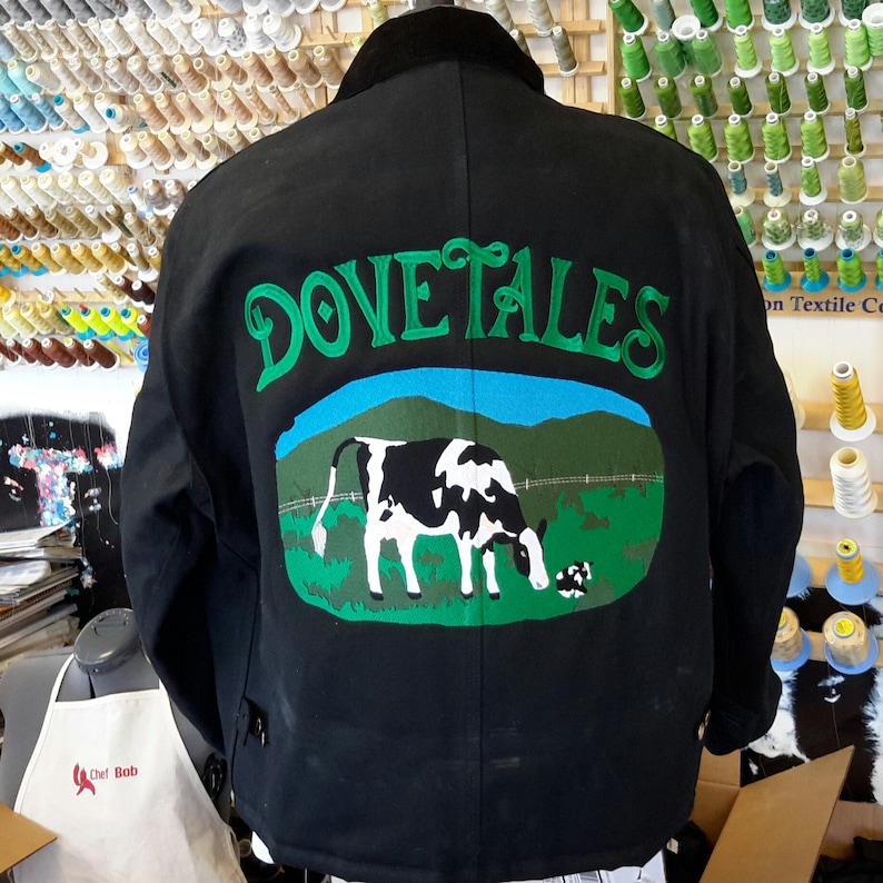 May include: A black work jacket with a green embroidered design of a cow in a field with the words "Dove Tales" above it.