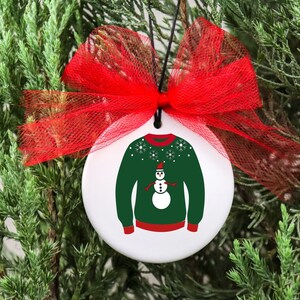 May include: A white ceramic Christmas ornament with a green sweater and a snowman design. The ornament has a red bow and a string for hanging.