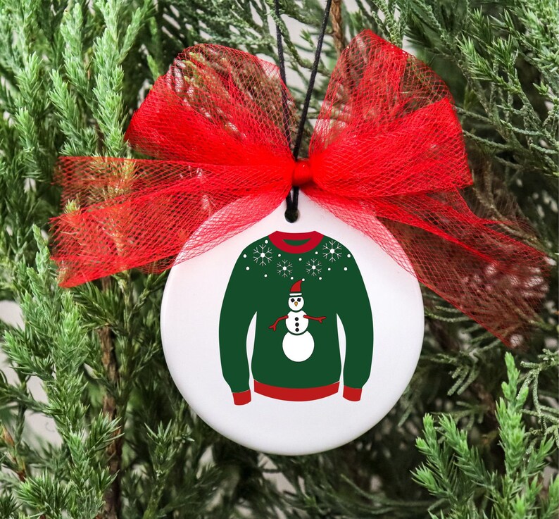 May include: A white ceramic Christmas ornament with a green sweater and a snowman design. The ornament has a red bow and a string for hanging.