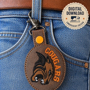 May include: A brown leather keychain with a metal clasp attached to blue jeans. The keychain features an embroidered cougar logo in black and orange, with the word "COUGARS" in orange. A digital download embroidery design is also visible.