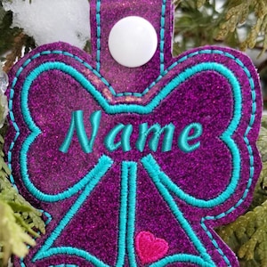 May include: Purple glitter fabric key chain with a turquoise bow and the word "Name" embroidered in the centre. A small pink heart is sewn at the bottom of the bow.