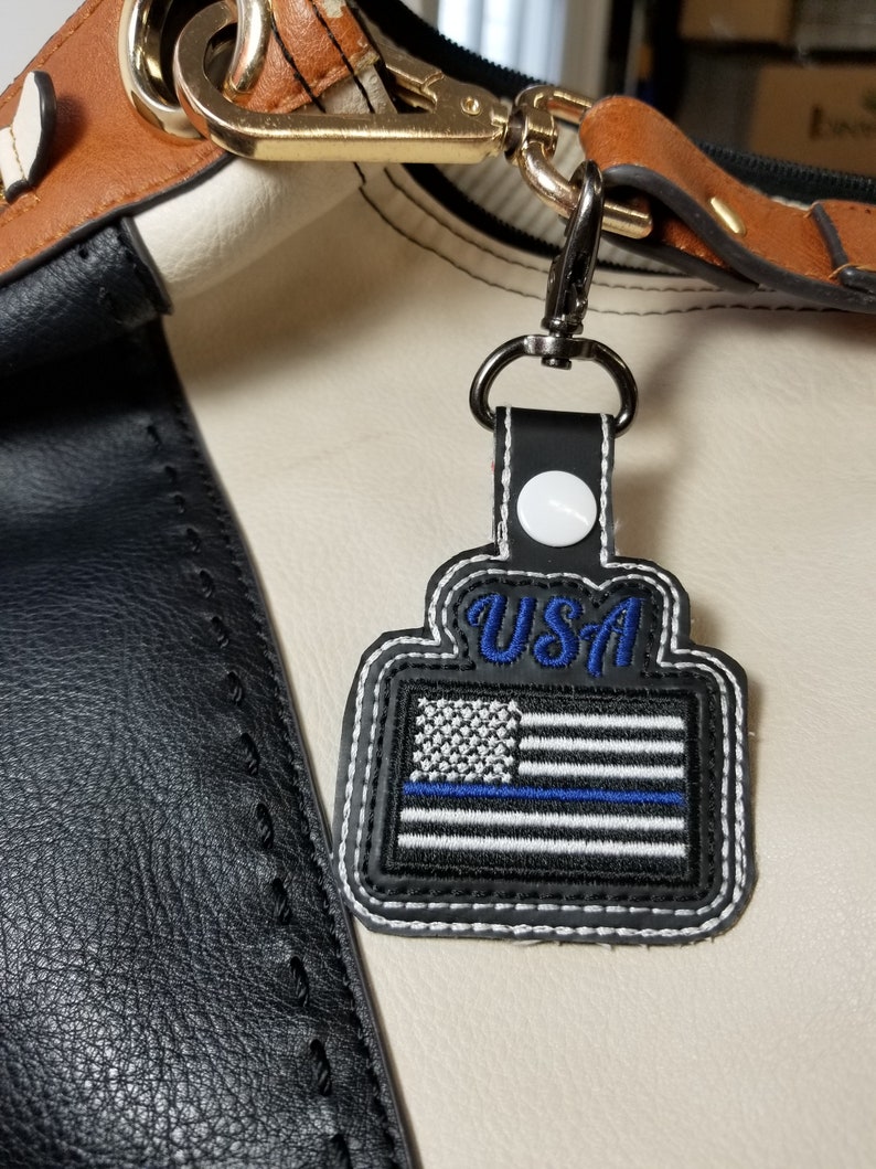 Puede incluir: Parche de la bandera de EE. UU. en blanco y negro con una l&iacute;nea azul, cosido sobre un fondo negro similar al cuero. La palabra "USA" est&aacute; bordada en azul. El parche est&aacute; sujeto a un bolso con un cierre plateado.