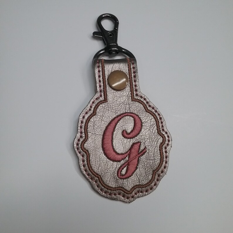 May include: A metallic silver keychain fob with a scalloped edge, featuring a large embroidered pink letter G in the center. The fob is secured with a bronze-colored snap button and attached to a black metal lobster claw clasp for easy attachment.