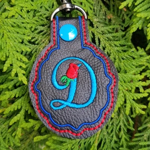 May include: A circular dark gray textured keychain featuring a bright blue embroidered letter D with a small red and green rose accent. The keychain has a blue snap button, red and blue decorative stitching, and a silver metal lobster clasp.