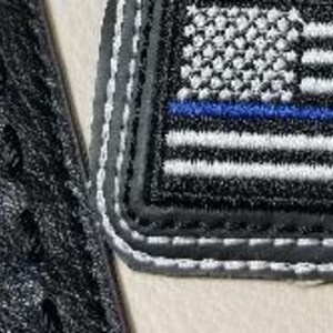 Puede incluir: Parche bordado con una bandera estadounidense en escala de grises con una l&iacute;nea azul, bordeado con costuras grises y blancas. El parche es rectangular y tiene un borde negro. Tambi&eacute;n se ve parte de una superficie negra texturizada.