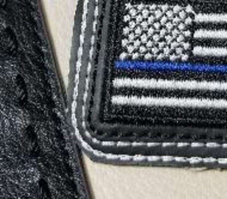 Puede incluir: Parche bordado con una bandera estadounidense en escala de grises con una l&iacute;nea azul, bordeado con costuras grises y blancas. El parche es rectangular y tiene un borde negro. Tambi&eacute;n se ve parte de una superficie negra texturizada.