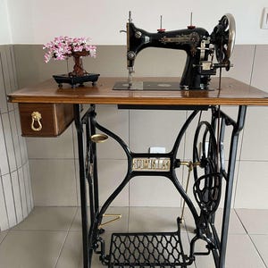 Könnte beinhalten: Eine antike schwarze Nähmaschine mit einer Holzplatte und einem dekorativen Bonsai-Baum. Die Maschine hat ein großes Handrad und ein Fußpedal. Der Tisch hat eine Schublade mit einem goldfarbenen Griff. Die Maschine steht auf einem schwarzen Metallrahmen.