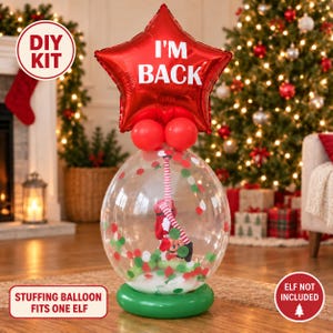 Op de afbeelding: Een doorzichtige ballon gevuld met rode, witte en groene confetti met een kleine elfpop, bekroond met een rode stervormige ballon met de witte tekst I'M BACK, rustend op een groene ronde basis. Bevat de tekst DIY KIT en ELF NOT INCLUDED.