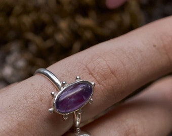 May include: A silver ring with a purple gemstone set in a starburst design. The ring is on a finger with a brown nail polish.