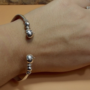 May include: A silver bangle bracelet with a series of round beads on the ends. The bracelet is open and has a simple design.