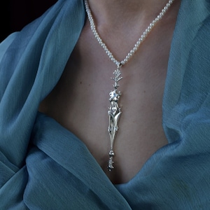 Peut inclure: Un collier en argent avec un pendentif représentant une figure féminine stylisée. Le collier est composé de petites perles blanches et est porté par une personne avec une écharpe bleue.