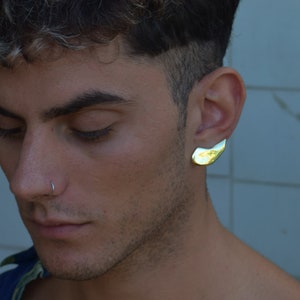 Op de afbeelding: Een close-up van een gezicht met kort, donker haar. De persoon draagt een neusring en een gouden oorring in de vorm van een halve maan.