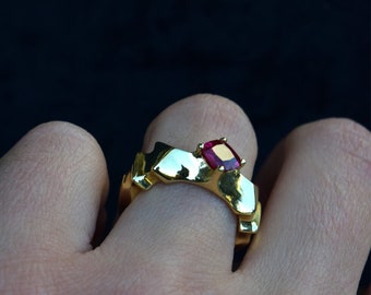 May include: A gold ring with a red gemstone set in the centre. The ring has a textured, geometric design.