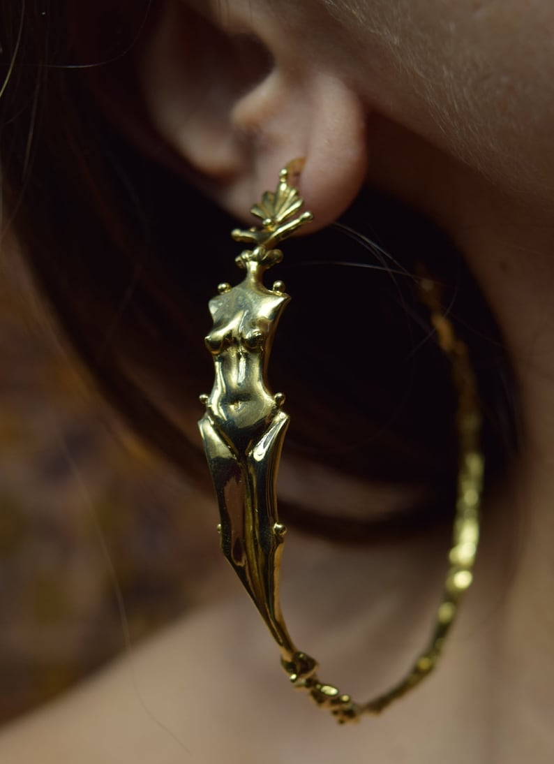 May include: Gold hoop earrings with a detailed figure of a woman's torso hanging from the hoop.