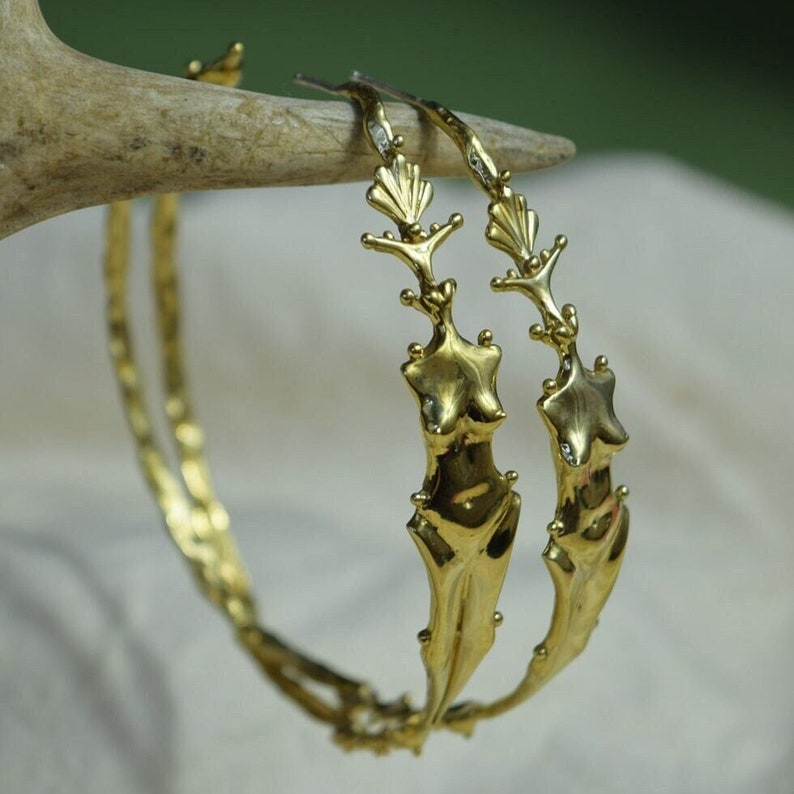 May include: A pair of gold hoop earrings with a detailed design of a stylized female figure. The earrings are made of a textured metal and have a slightly hammered finish.