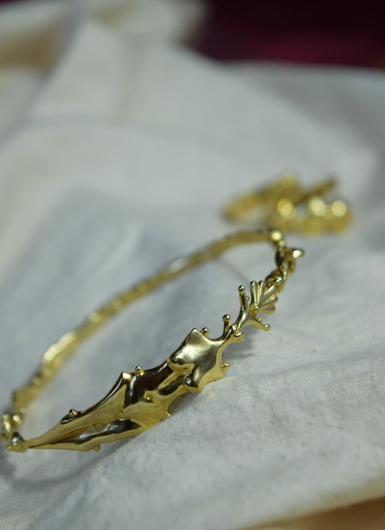 May include: A gold-toned bangle bracelet with a unique, organic design. The bracelet is made of twisted metal and features a large, abstract shape that resembles a branch or a piece of coral.