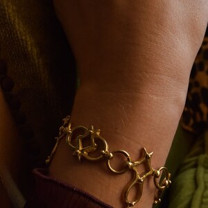 Puede incluir: Un brazalete dorado con un dise&ntilde;o &uacute;nico e intrincado que presenta c&iacute;rculos entrelazados y p&uacute;as. El brazalete se lleva en la mu&ntilde;eca.