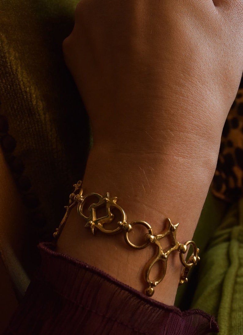 Puede incluir: Un brazalete dorado con un dise&ntilde;o &uacute;nico e intrincado que presenta c&iacute;rculos entrelazados y p&uacute;as. El brazalete se lleva en la mu&ntilde;eca.