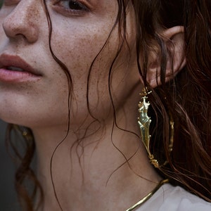 May include: A woman with freckles and wet hair wears a gold necklace and a large gold hoop earring with a textured design.