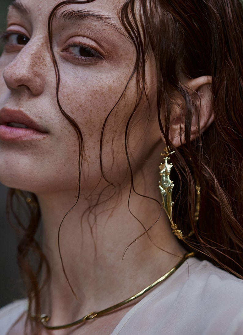 May include: A woman with freckles and wet hair wears a gold necklace and a large gold hoop earring with a textured design.