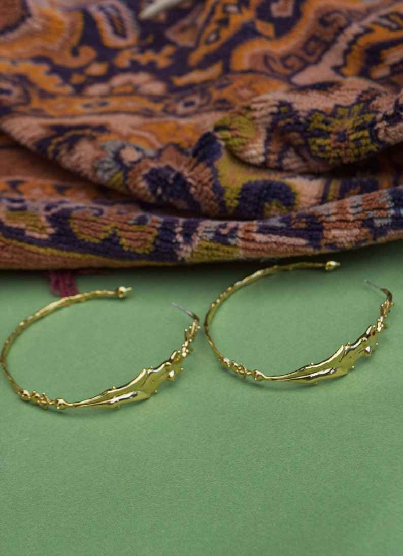 May include: A pair of gold hoop earrings with a unique, abstract design. The earrings are made of a thick, textured metal and have a slightly irregular shape.