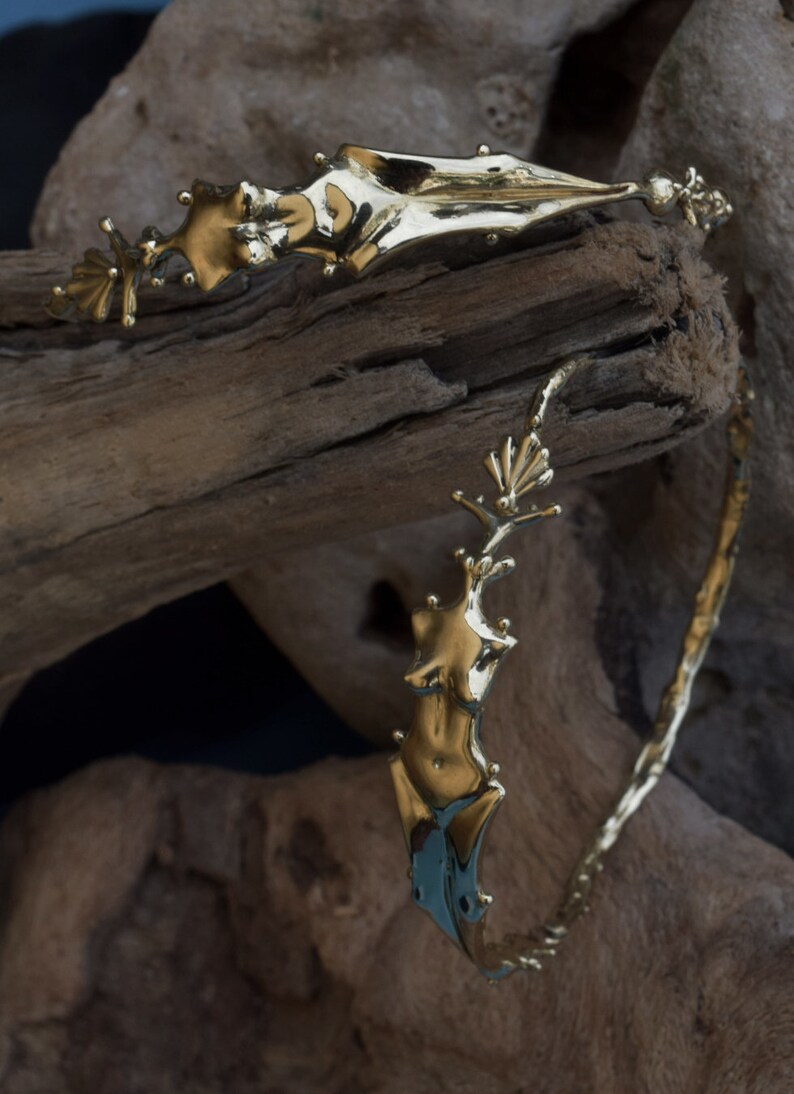 May include: A gold-toned metal bracelet with a unique design featuring a stylized human figure. The bracelet is crafted with a textured, organic-looking surface.