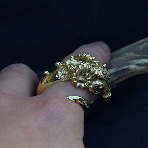 May include: A gold ring featuring a detailed ram's head design. The ring is worn on a person's finger.