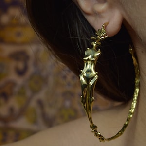 May include: Gold hoop earrings with a detailed figure of a woman's body in the center.