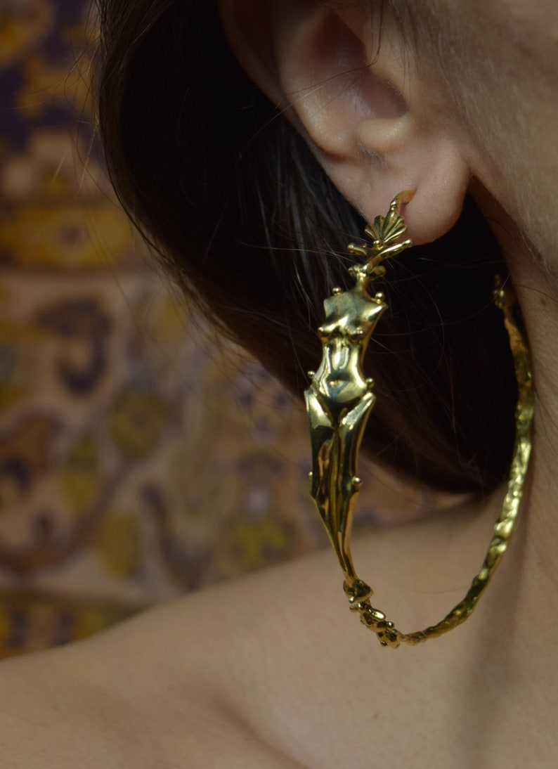 May include: Gold hoop earrings with a detailed figure of a woman's body in the center.