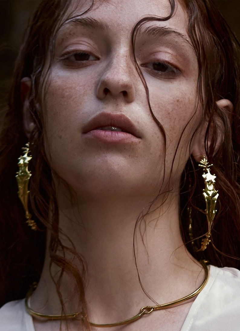 May include: A woman with wet, brown hair wears a gold choker necklace and large, gold, ornate earrings.