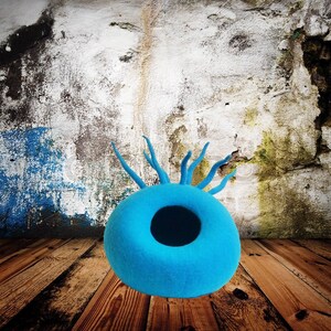 May include: A bright blue felt cat bed shaped like a donut with a hole in the center. The bed has a textured surface and is sitting on a wooden floor.