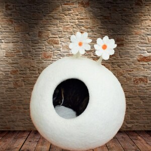 May include: White felt cat bed shaped like a sphere with two white felt flowers on top. The bed has a round opening for the cat to enter.
