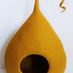 May include: A mustard yellow felt cat bed shaped like a teardrop with a curled felt tail. The bed has a round opening for the cat to enter.