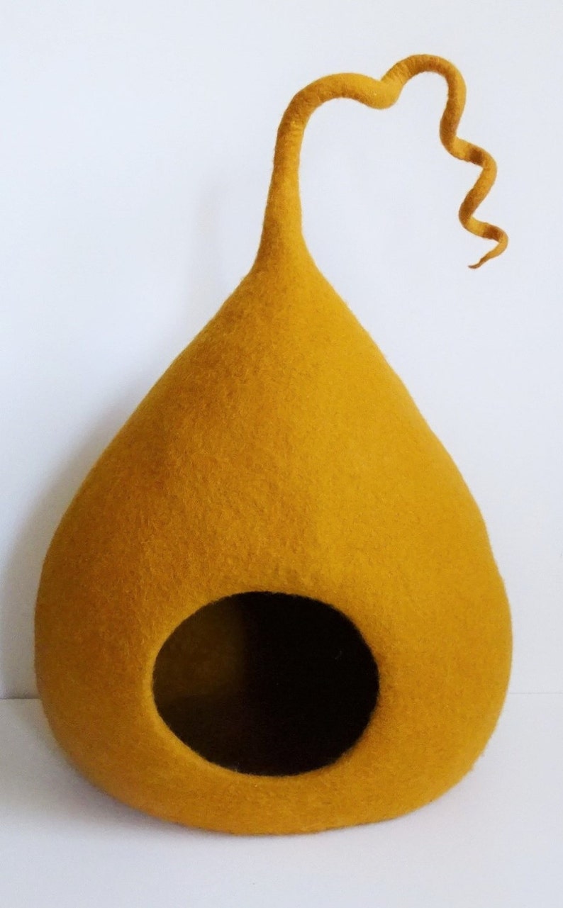 May include: A mustard yellow felt cat bed shaped like a teardrop with a curled felt tail. The bed has a round opening for the cat to enter.