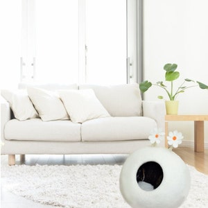 May include: White felt cat bed shaped like a sphere with a round entrance hole. The bed has two white felt flowers with orange centers on top. The bed is on a white shag rug in front of a white sofa.