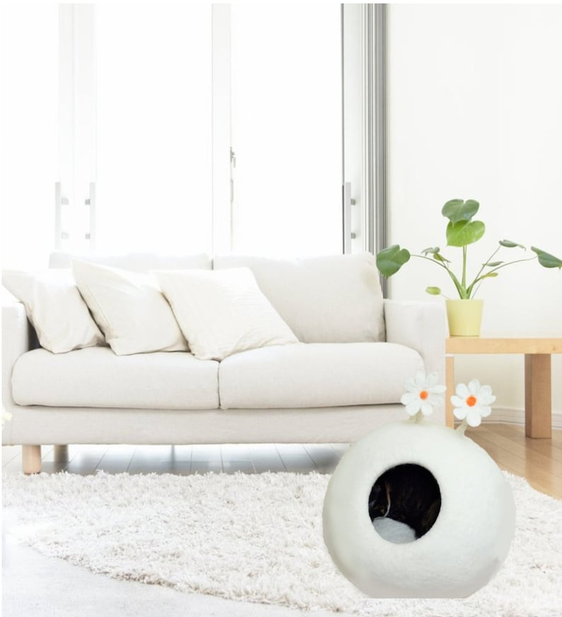 May include: White felt cat bed shaped like a sphere with a round entrance hole. The bed has two white felt flowers with orange centers on top. The bed is on a white shag rug in front of a white sofa.
