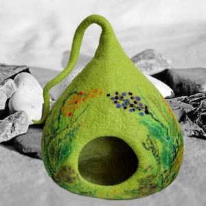 Puede incluir: Una cama para mascotas de fieltro verde con forma de lágrima con una pequeña abertura para la entrada. La cama tiene un exterior de fieltro verde con diseños florales pintados en tonos naranja, azul y verde.