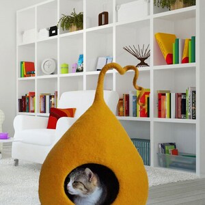 May include: A mustard yellow felt cat bed shaped like a teardrop with a curled felt tail. A gray and white cat is inside the bed.
