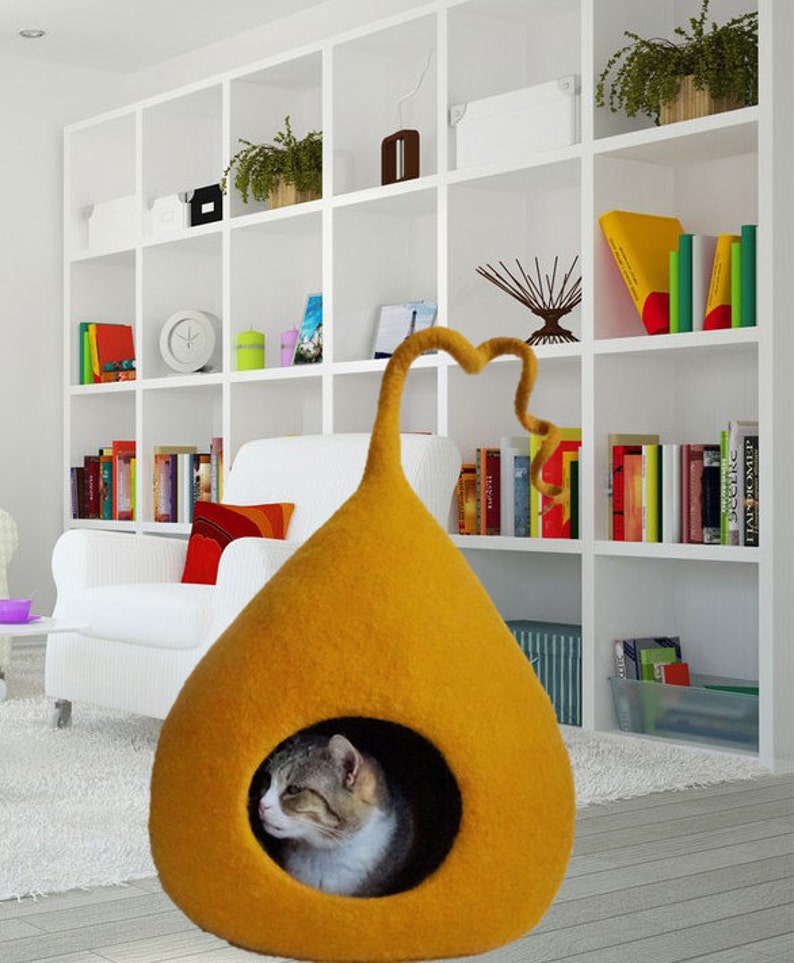May include: A mustard yellow felt cat bed shaped like a teardrop with a curled felt tail. A gray and white cat is inside the bed.