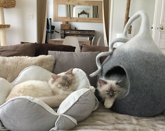 以下が含まれることがあります： 白とグレーの猫が2匹、グレーとホワイトの猫用ベッドで休んでいます。1匹の猫はグレーとホワイトの花の形をしたベッドで寝ており、もう1匹の猫はグレーとホワイトのフェルト製の猫用洞窟に座っています。